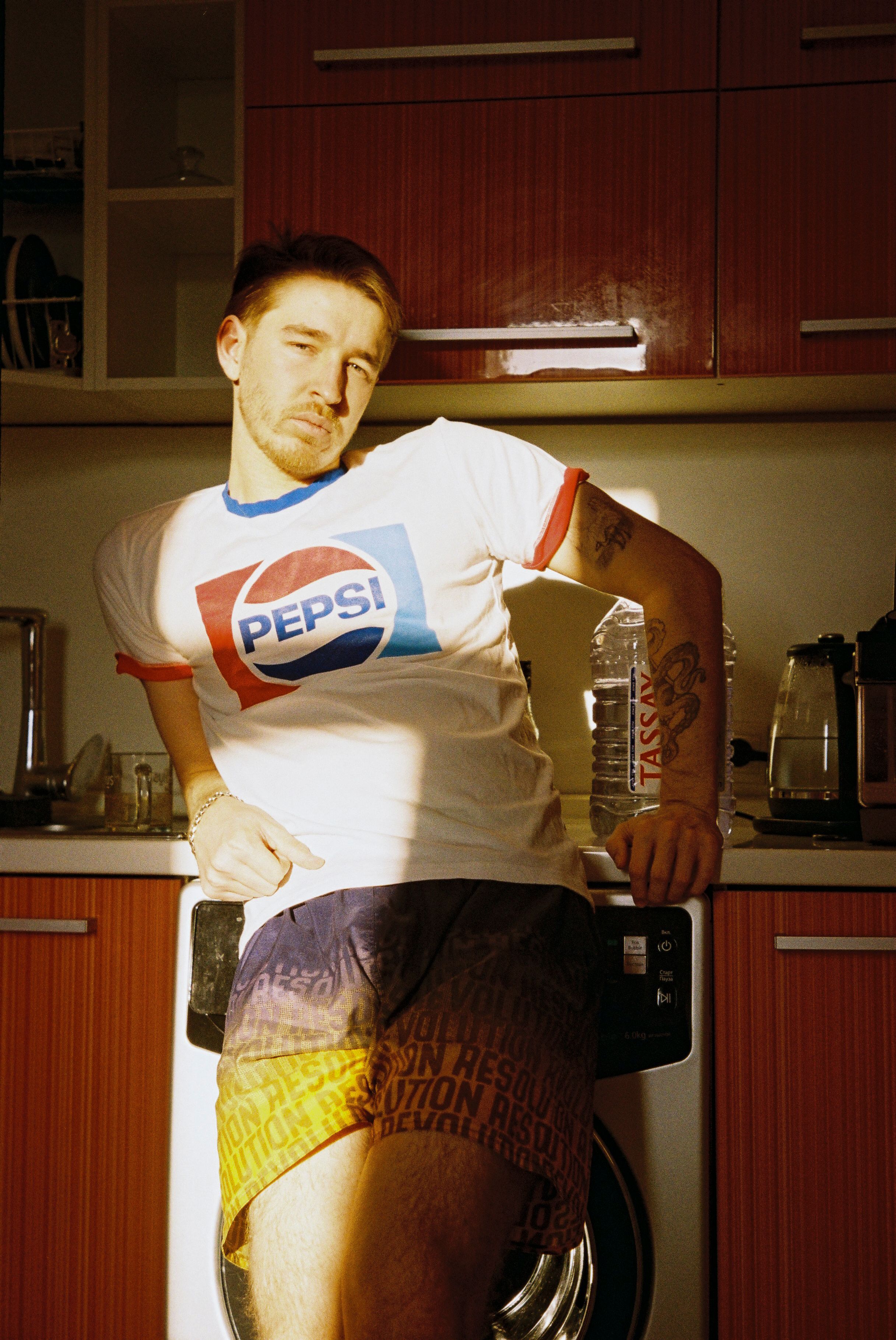 A relaxed portrait of a man with tattoos in a brightly lit kitchen wearing a Pepsi T-shirt, embracing vintage style.