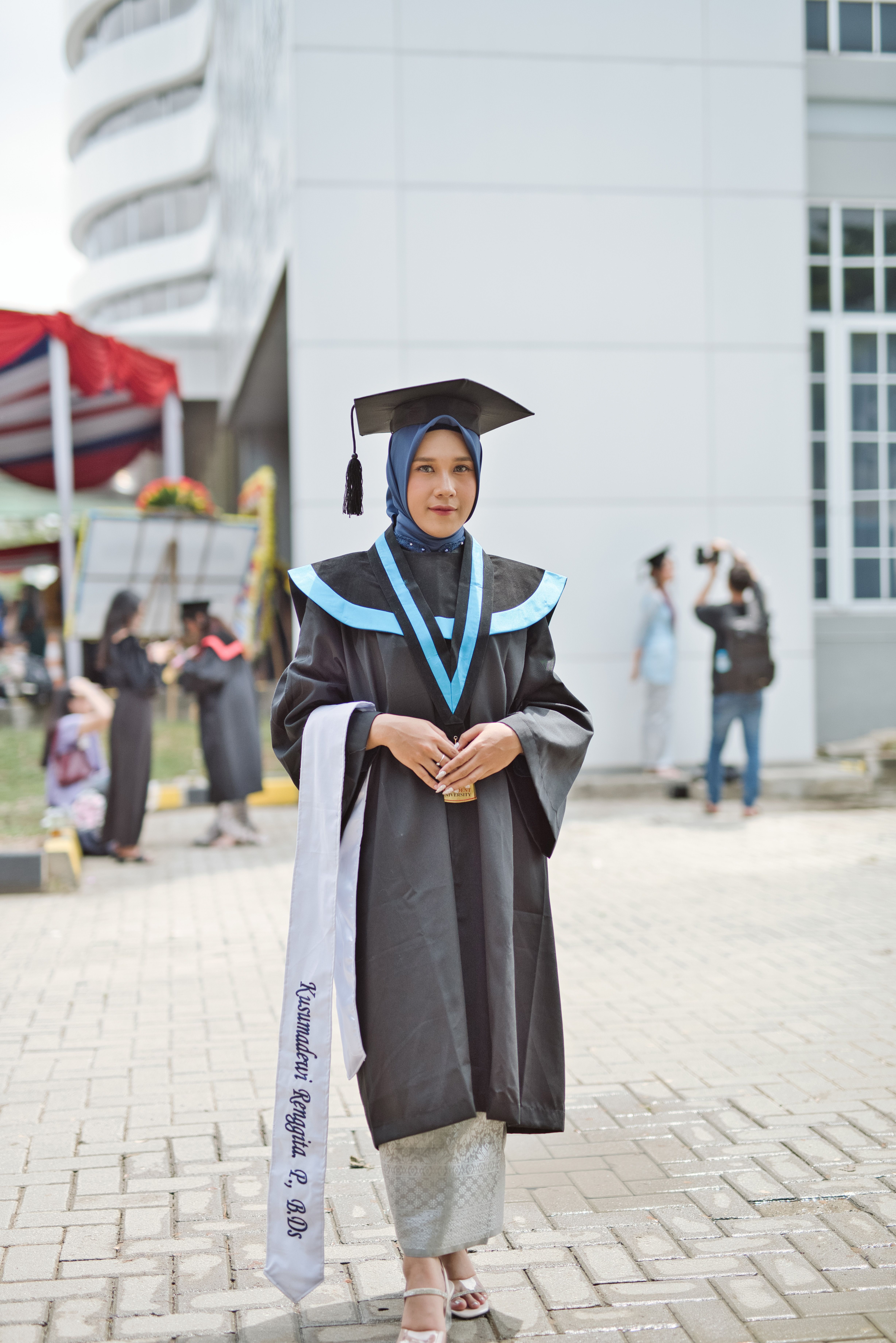 Female graduate in cap and gown at outdoor ceremony, holding diploma.