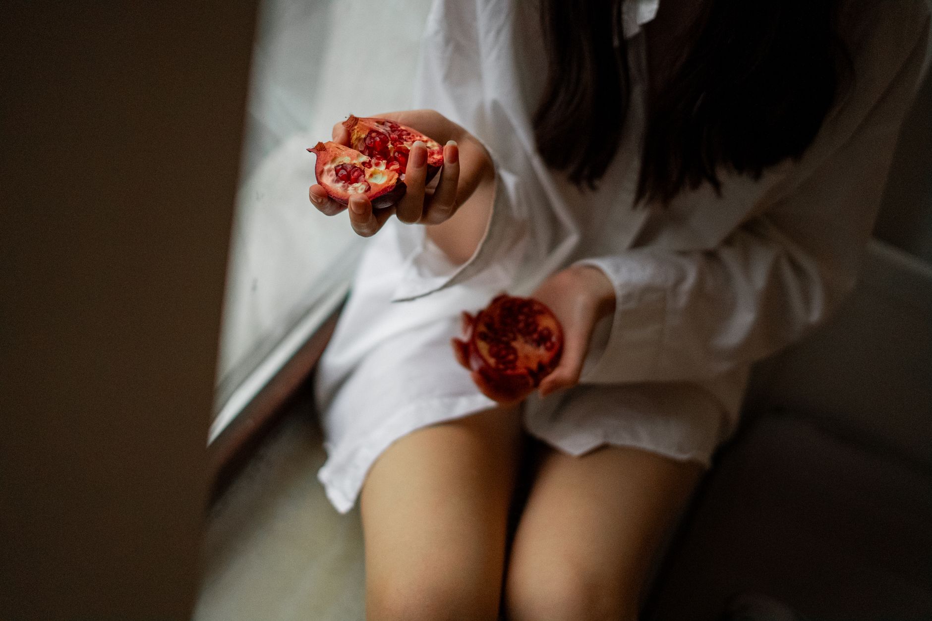 A young woman in casual attire holding a pomegranate by a window, capturing a serene moment.