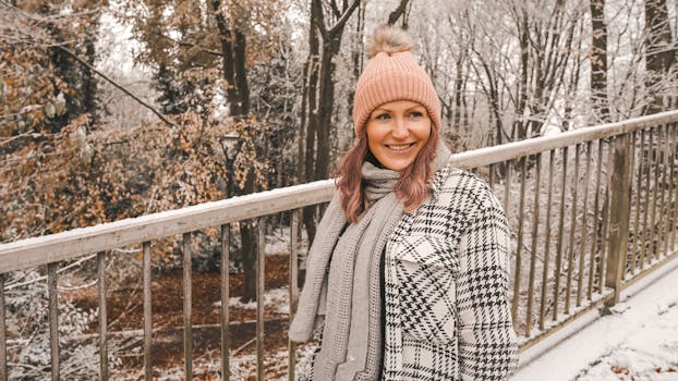 A smiling woman in winter attire stands on a snowy bridge in Cheadle, England, enjoying the winter scenery.