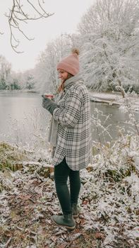 Woman holding a hot drink, standing by a frozen lake in winter, surrounded by snow.