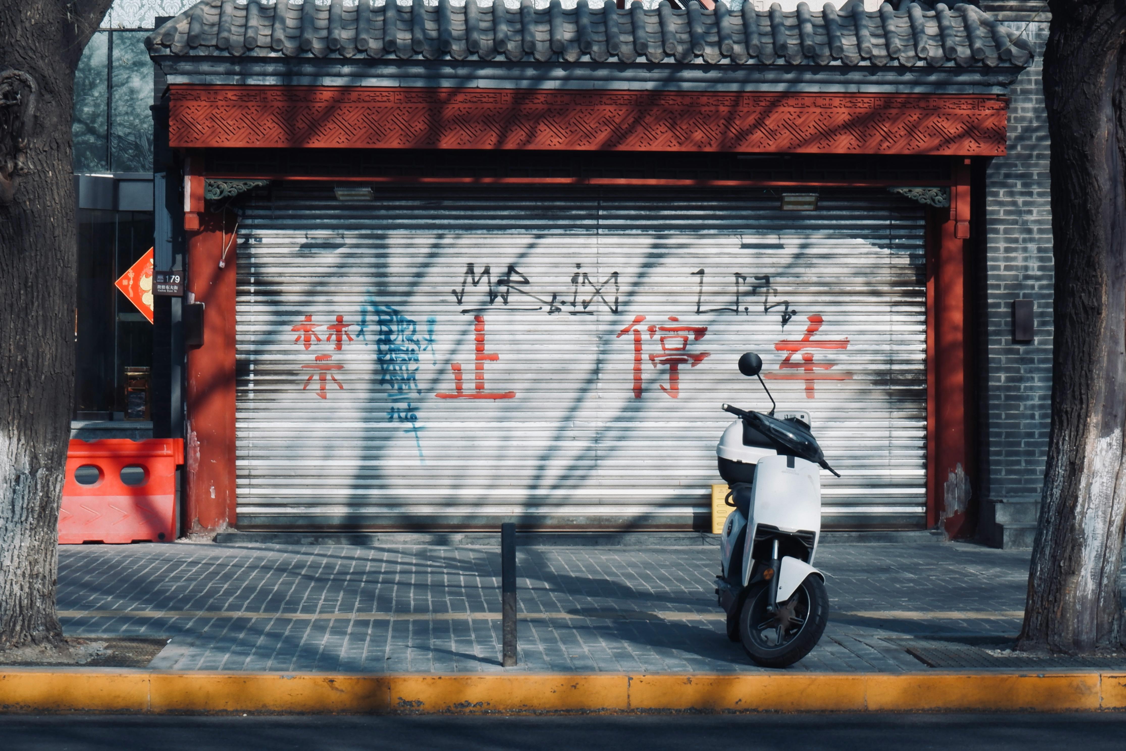 Ücretsiz Güneşli bir şehir sokağında, kepenkleri kapalı, grafitilerle kaplı bir dükkânın önüne park edilmiş bir scooter. Stok Fotoğraflar