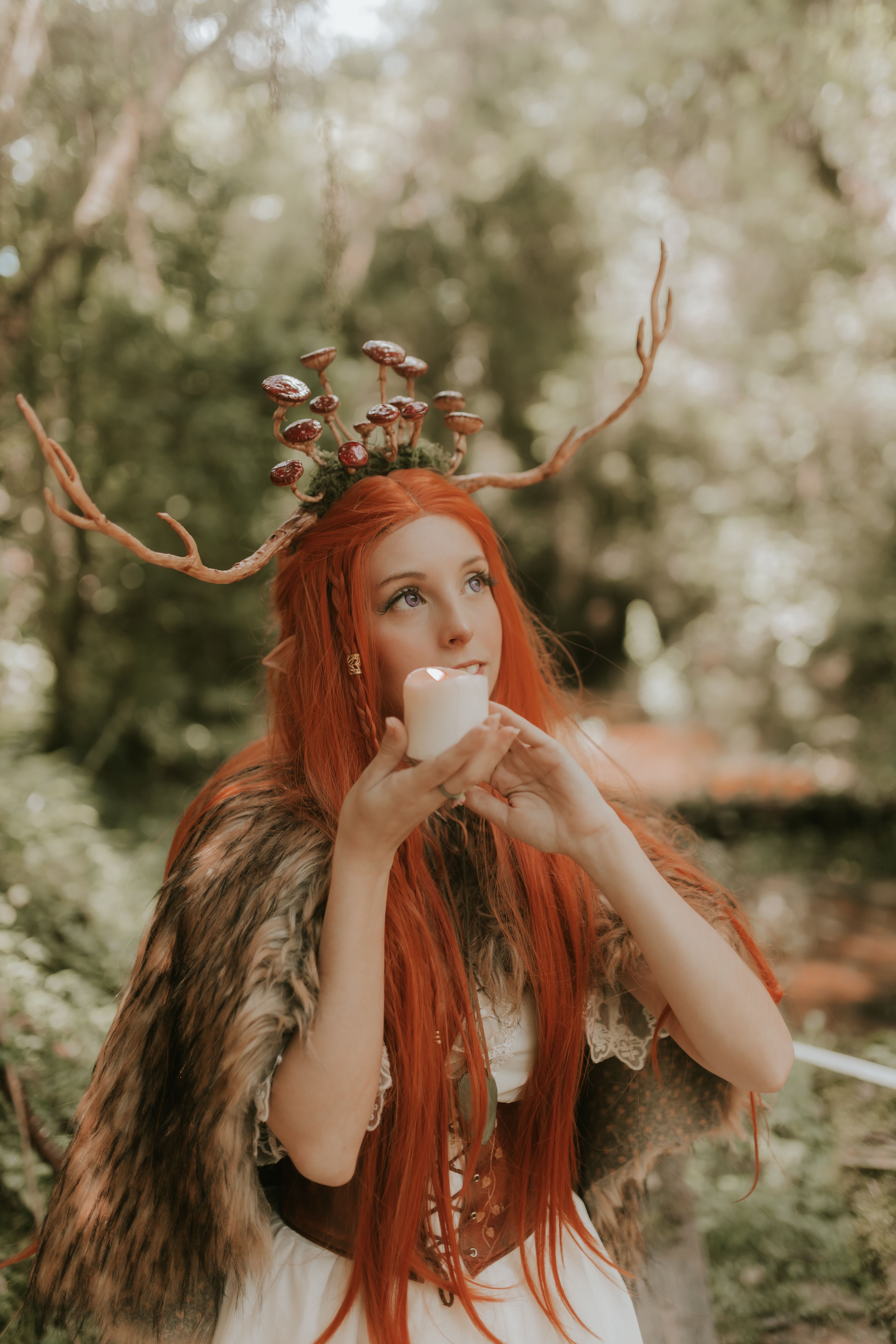 Mystical woman with red hair in a forest, wearing antlers and fur, holding a candle.