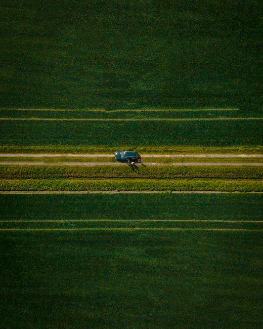 Drone shot capturing a car on a rural road surrounded by expansive green fields in Venlo, Netherlands.