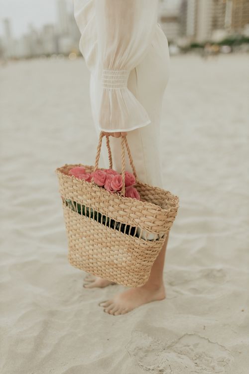 Free Elegant woman standing on a sandy beach holding a woven bag filled with roses. Stock Photo