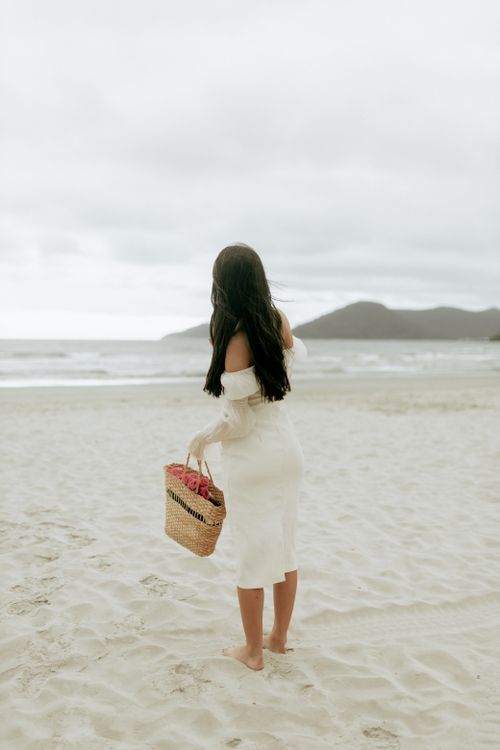 Free A woman in a white dress holding a woven basket on a tranquil beach, evoking serenity and relaxation. Stock Photo