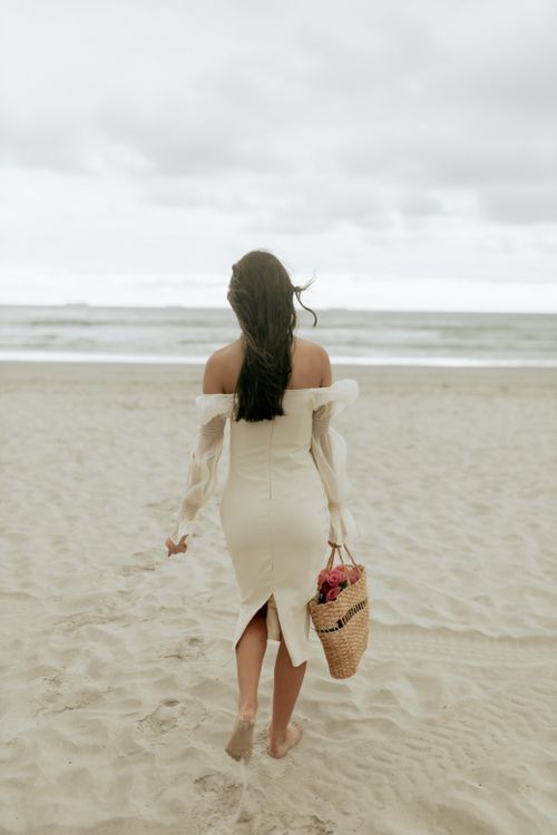 Free Woman in white dress strolling on a serene beach, carrying a straw bag. Stock Photo