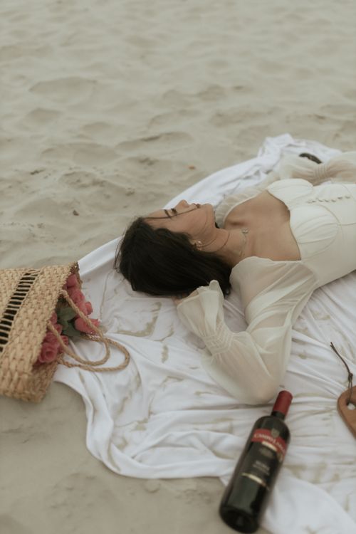 Free Serene beach scene with a woman lying on sand, wine bottle, and bouquet. Stock Photo