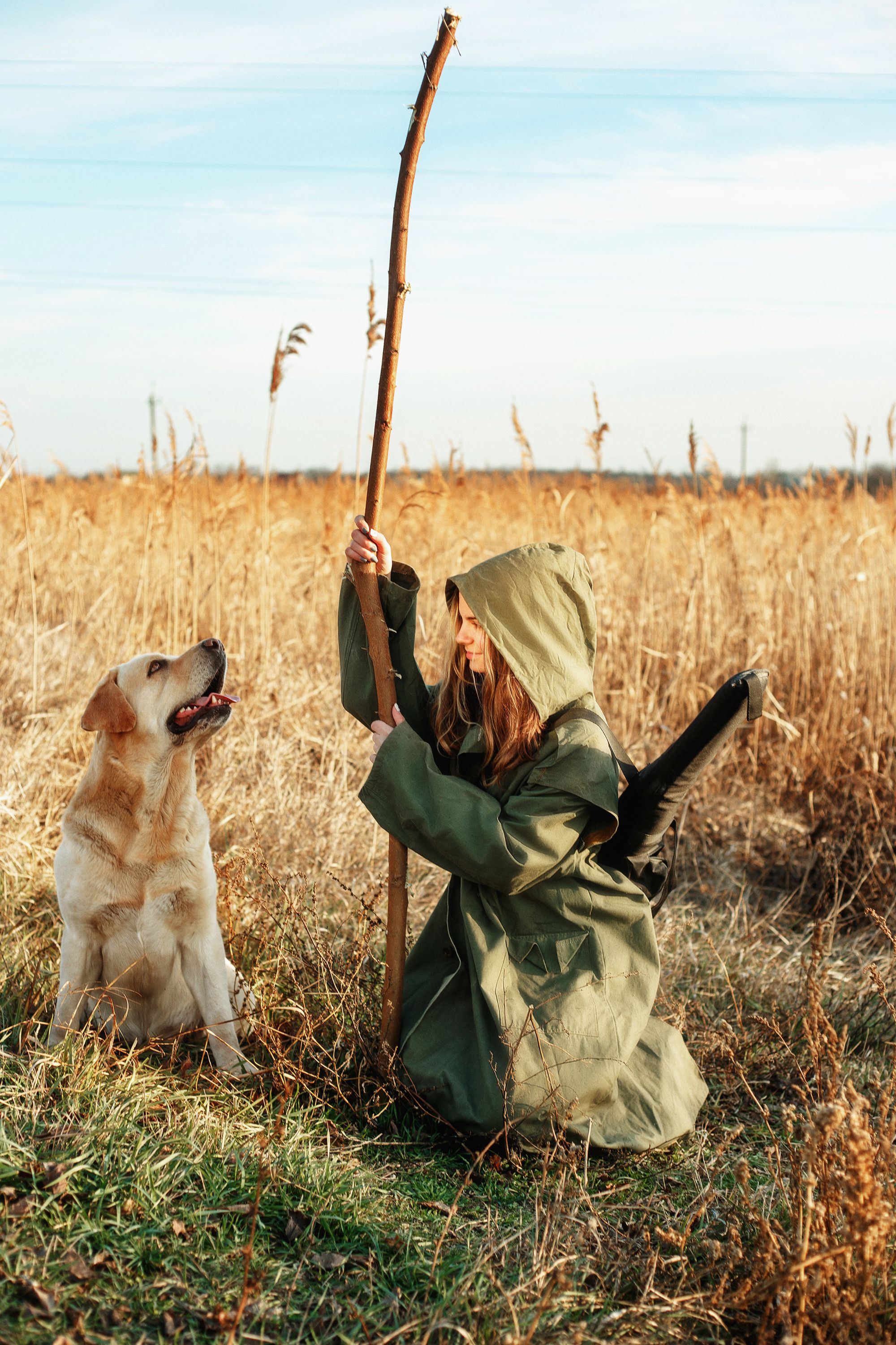 A young girl in a green coat with a dog in a rural field holding a stick.