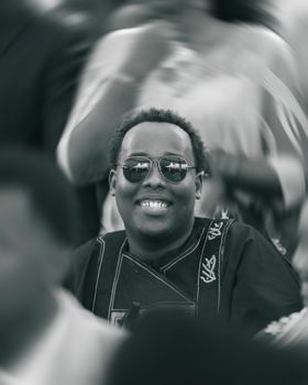 A cheerful man in sunglasses at an event, wearing traditional attire. Captured in a dynamic, crowded setting.