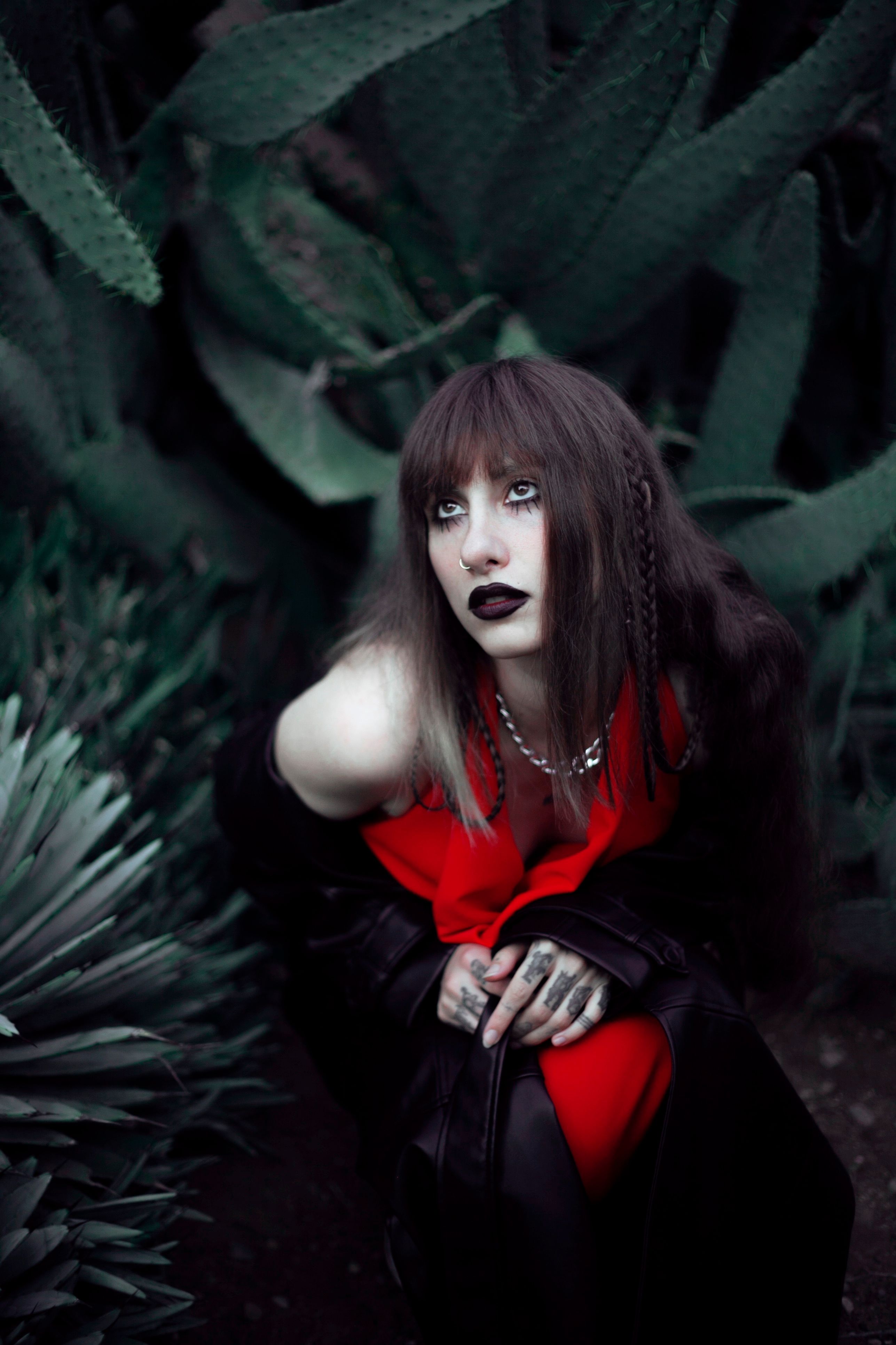 Artistic portrait of a gothic woman in red among lush dark green plants, conveying mystery.
