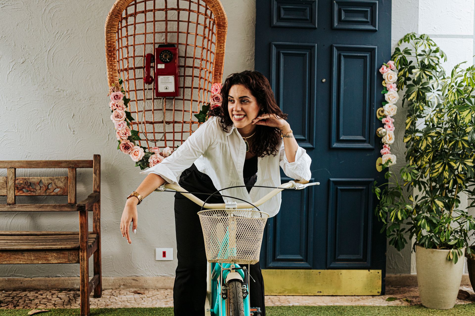 Smiling woman leans on a bicycle indoors, surrounded by floral decor and a vintage phone backdrop.