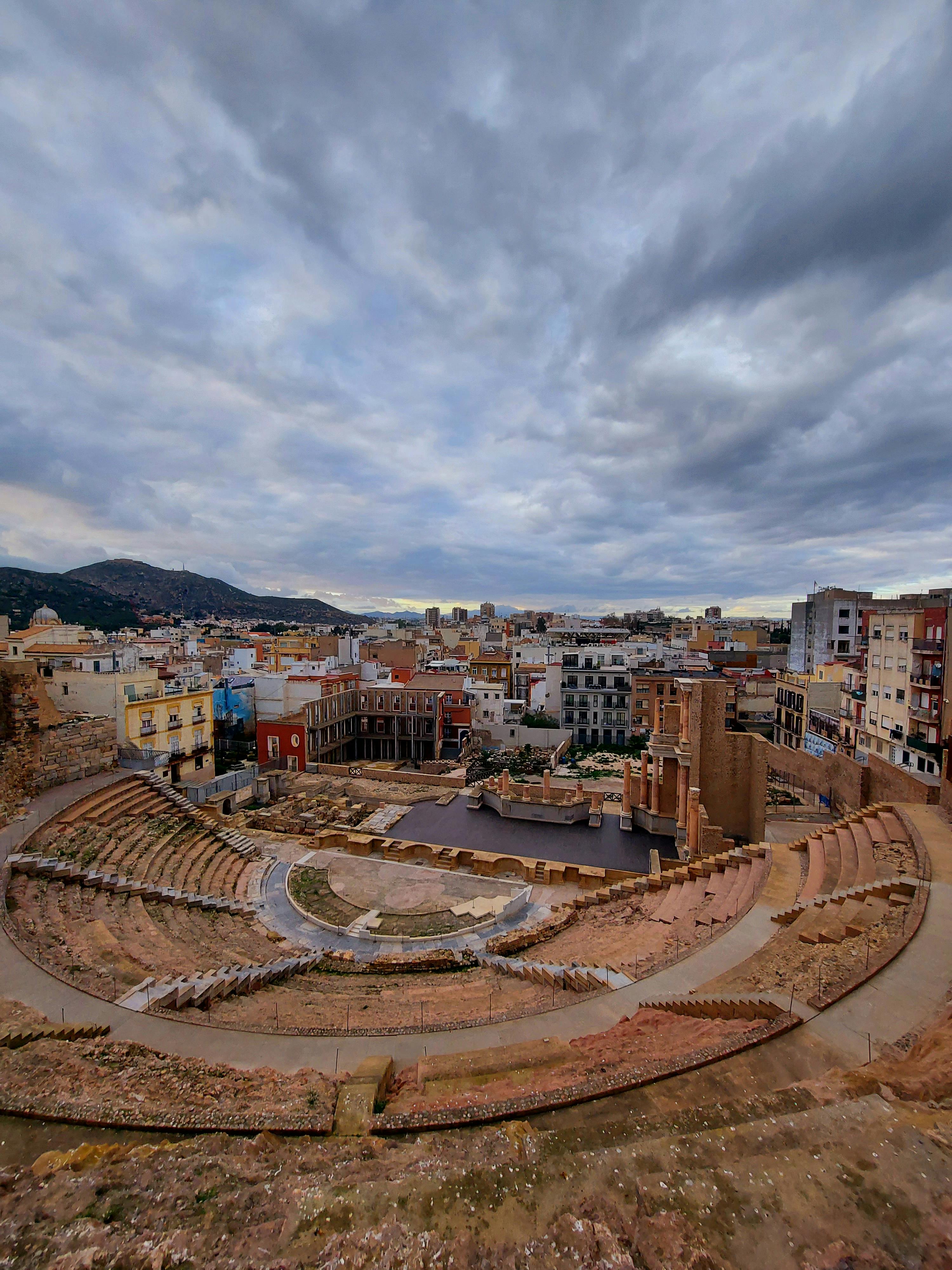 ingyenes Lenyűgöző kilátás egy ókori római amfiteátrumra, amely egy modern városkép közepette fészkelődik a drámai égbolt alatt. Stockfotó