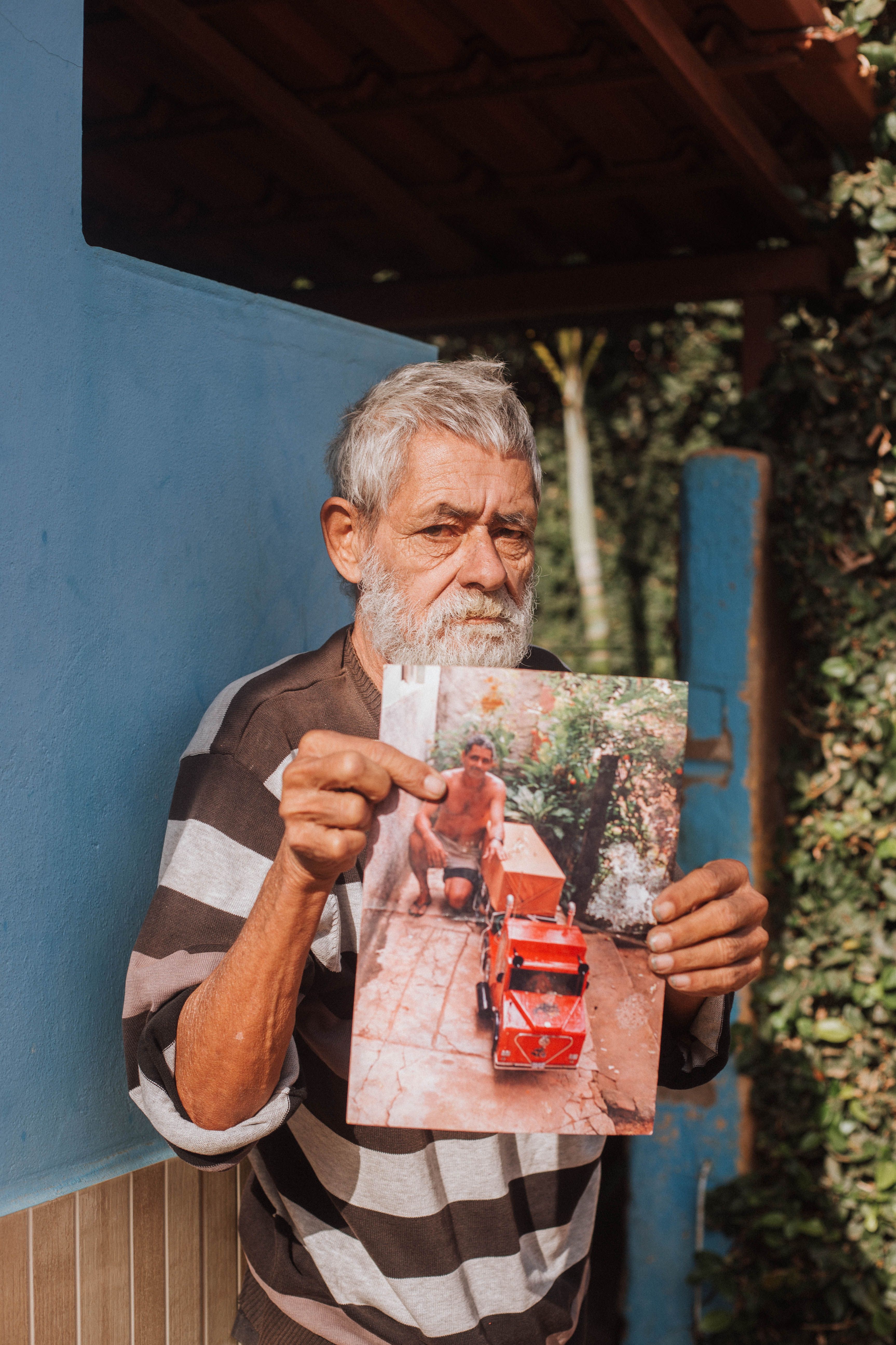 Elderly man in striped shirt holds a nostalgic photo outdoors, showcasing personal history.