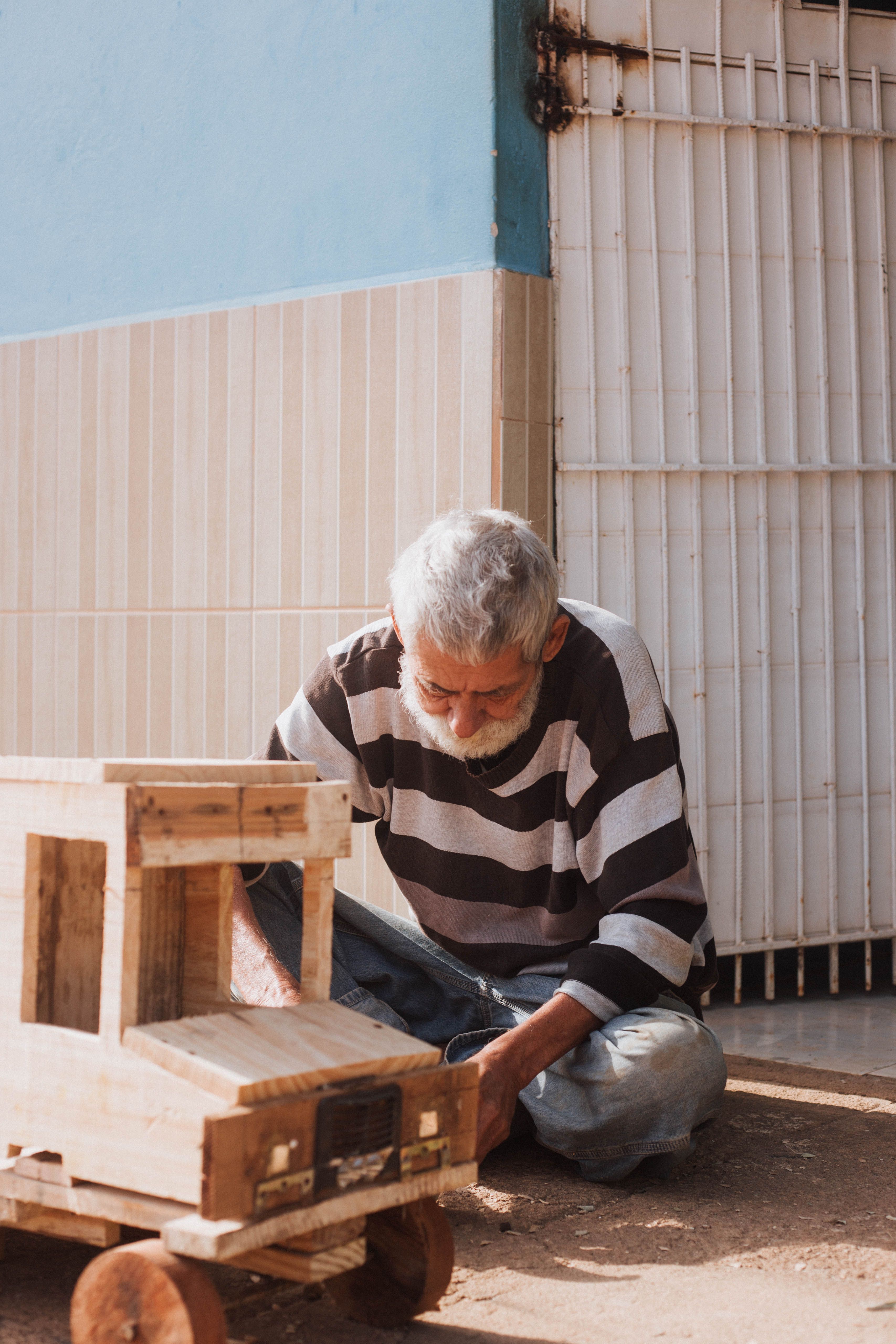 A senior man crafting with wood, seated on the ground in Tarumirim, Brazil.
