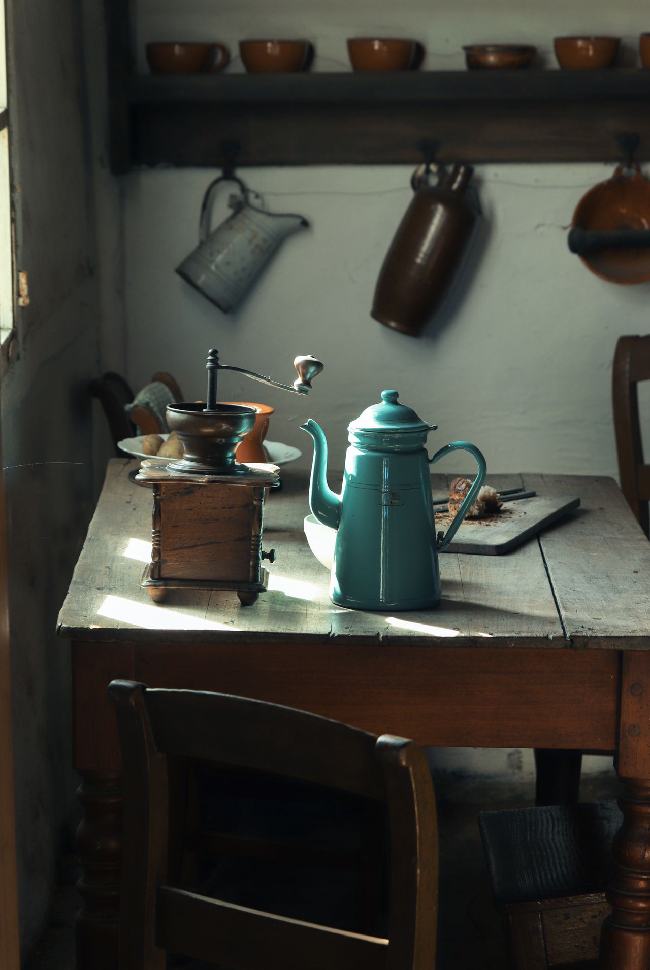 Old farmhouse kitchen with a sturdy wooden table, an enamel coffee pot, a vintage coffee grinder and bowls on open shelves