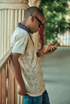 Man wearing sunglasses checks phone while leaning on railing outdoors.