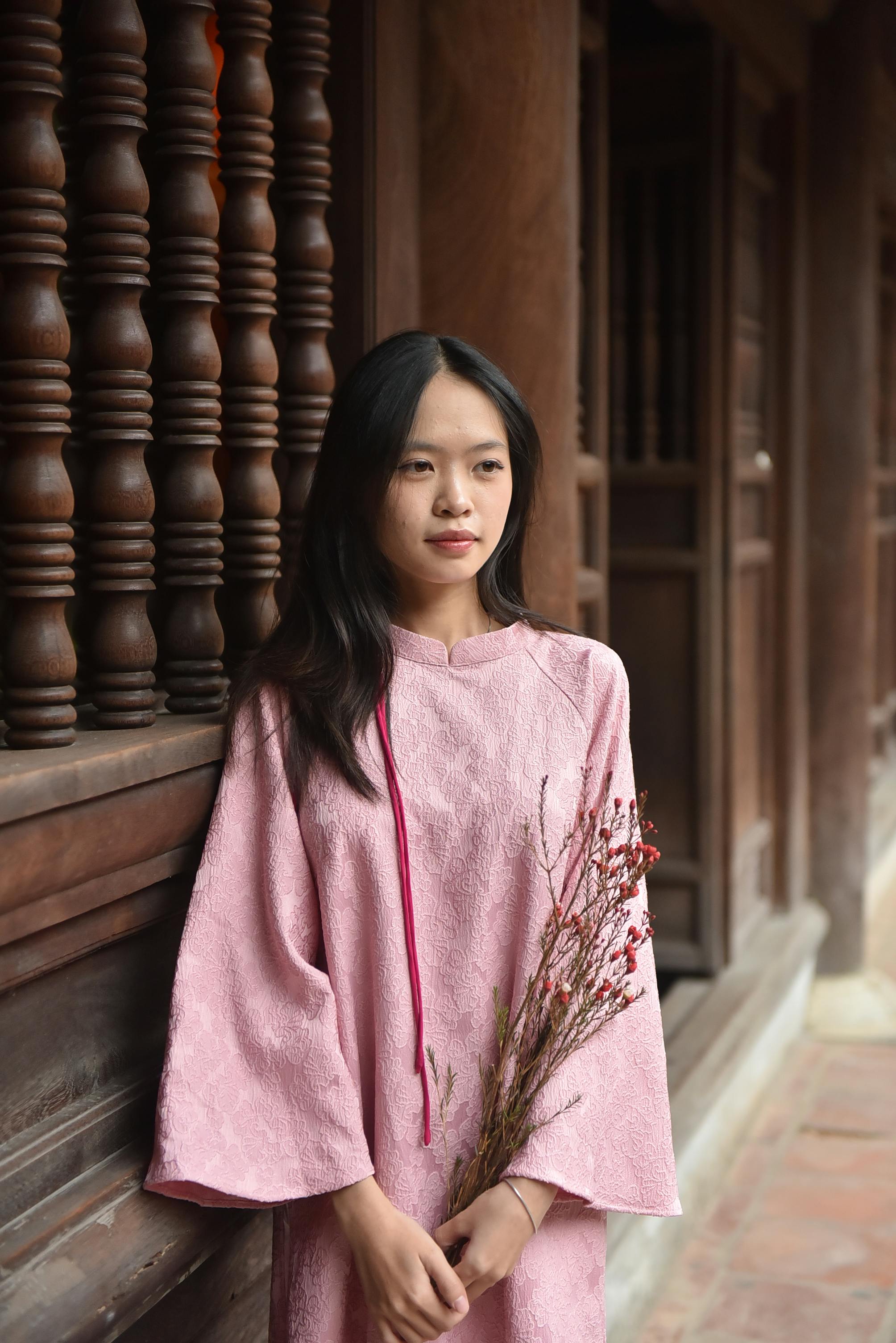 A young woman in a pink traditional dress holds flowers indoors, showcasing cultural elegance.