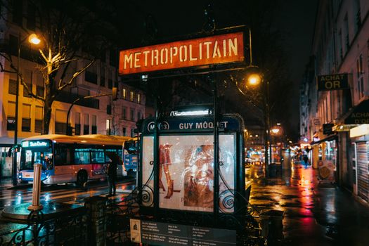 A wet Paris street captures the glow of city lights by the Metropolitain entrance at Guy Môquet metro station.