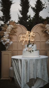 Beautiful wedding cake on elegant table with floral decor and neon sign backdrop outdoors.