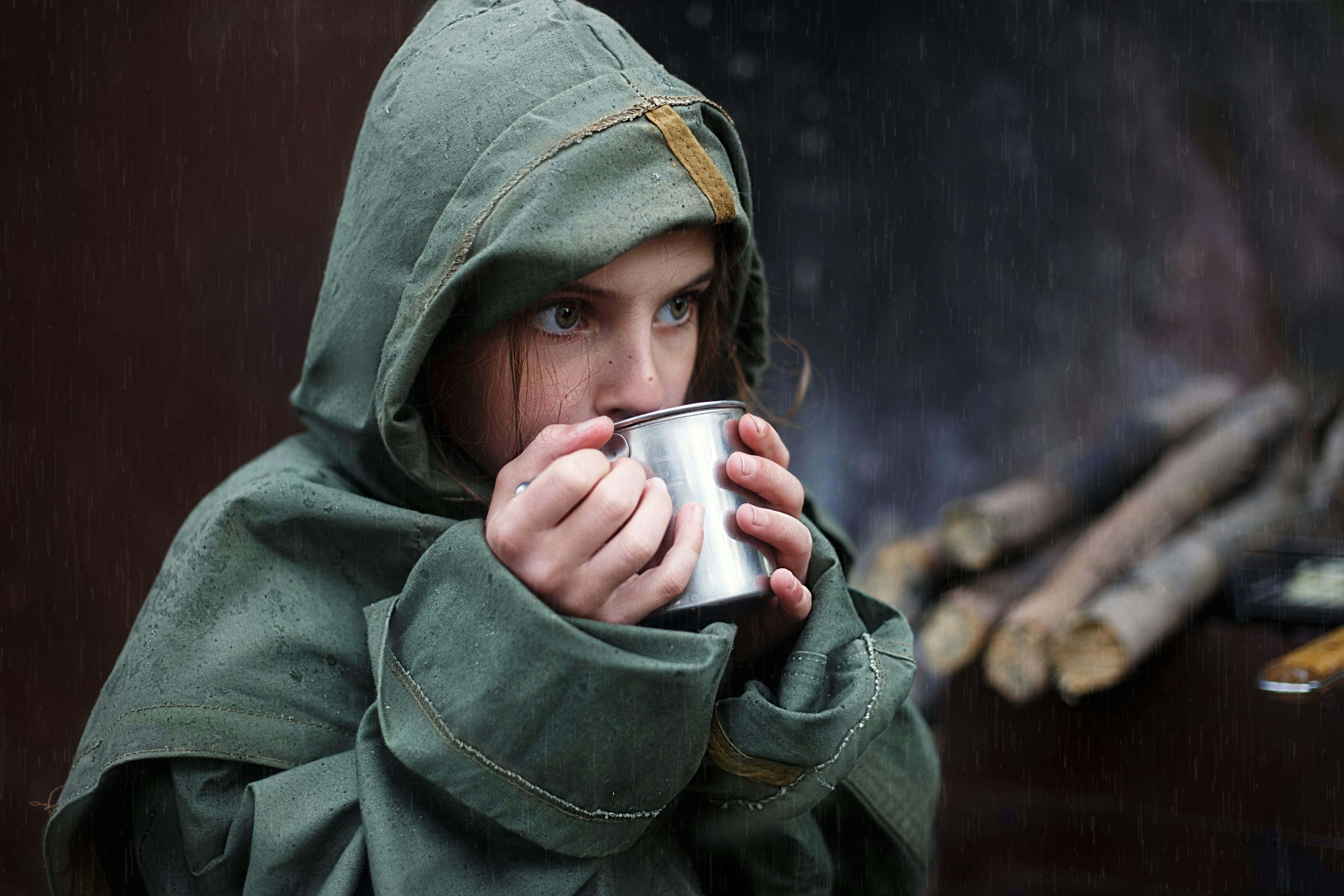 Young girl in raincoat sipping warm beverage on a rainy day in the forest.