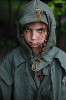 A young girl in a green raincoat with a serious expression stands outdoors in the rain.