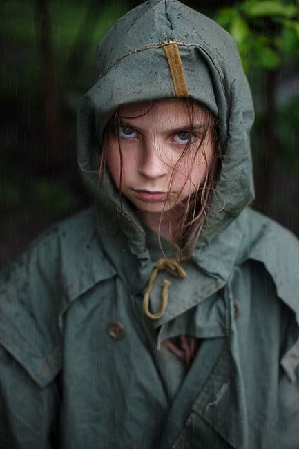 A young girl in a green raincoat with a serious expression stands outdoors in the rain.