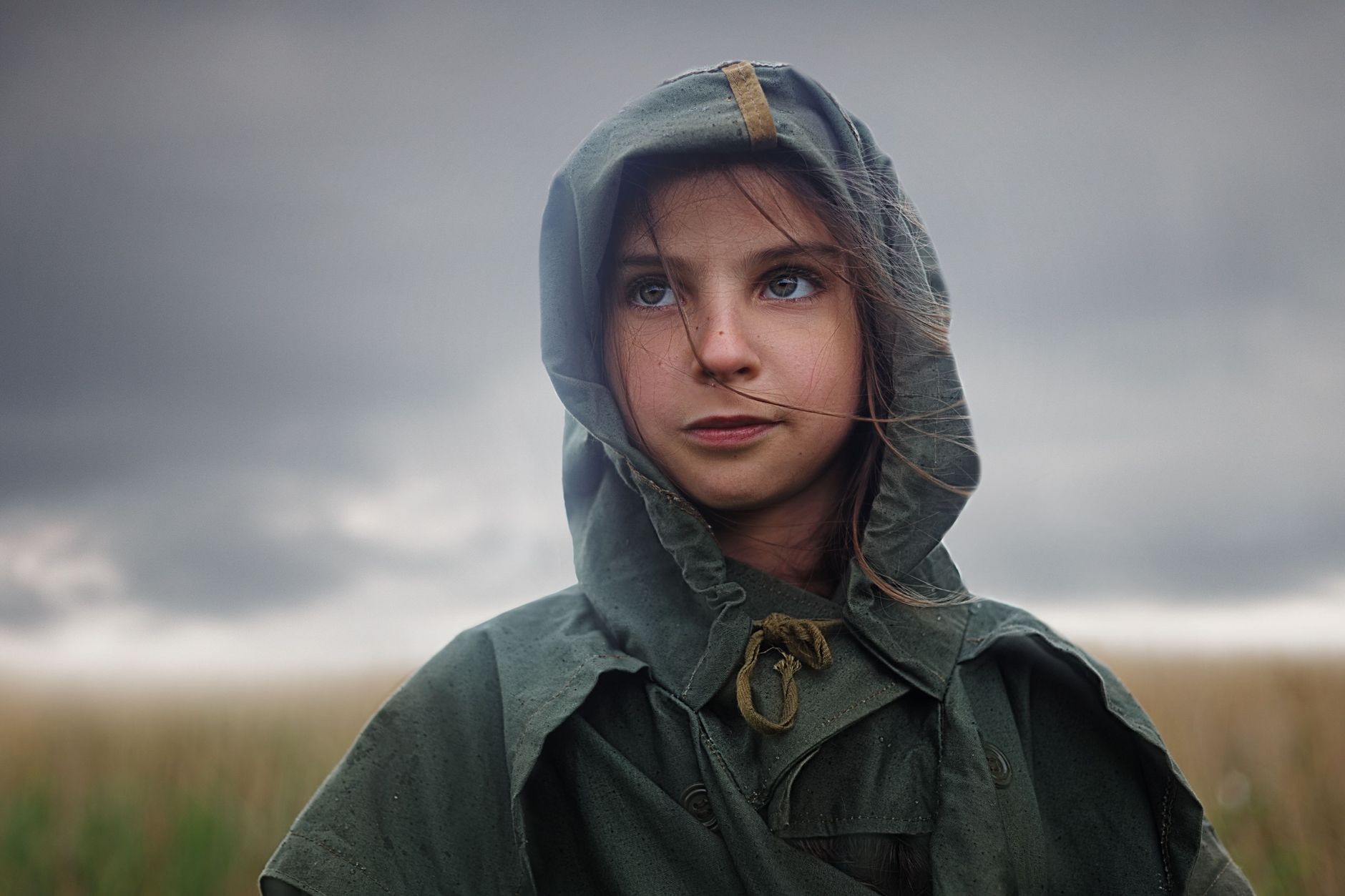 Portrait of a young woman in a hooded coat standing outdoors with a stormy sky backdrop.