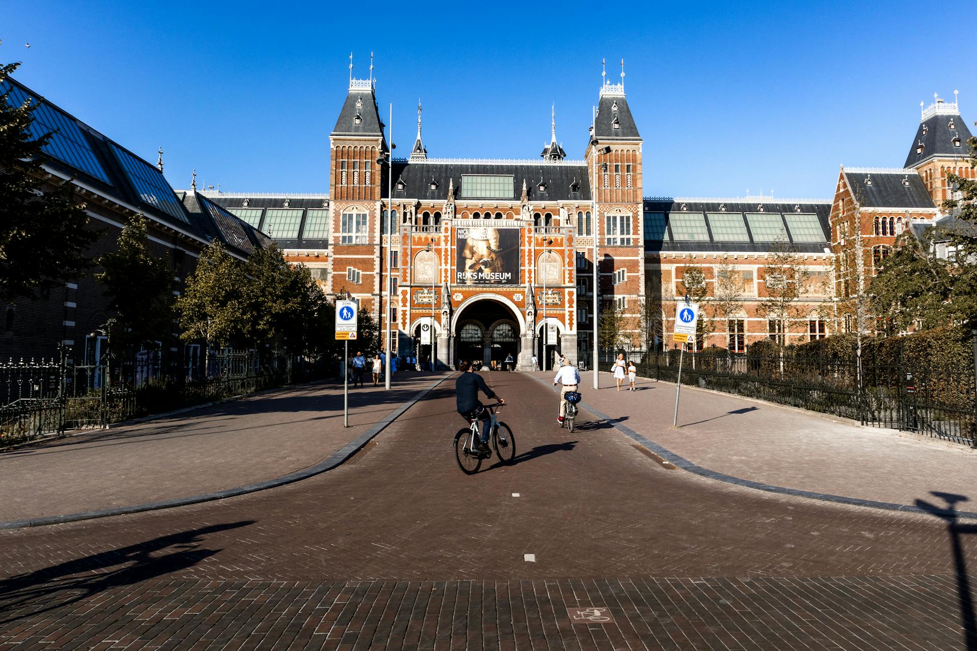 Cyclists ride past the iconic Rijksmuseum entrance in Amsterdam on a clear day, showcasing Dutch architecture.