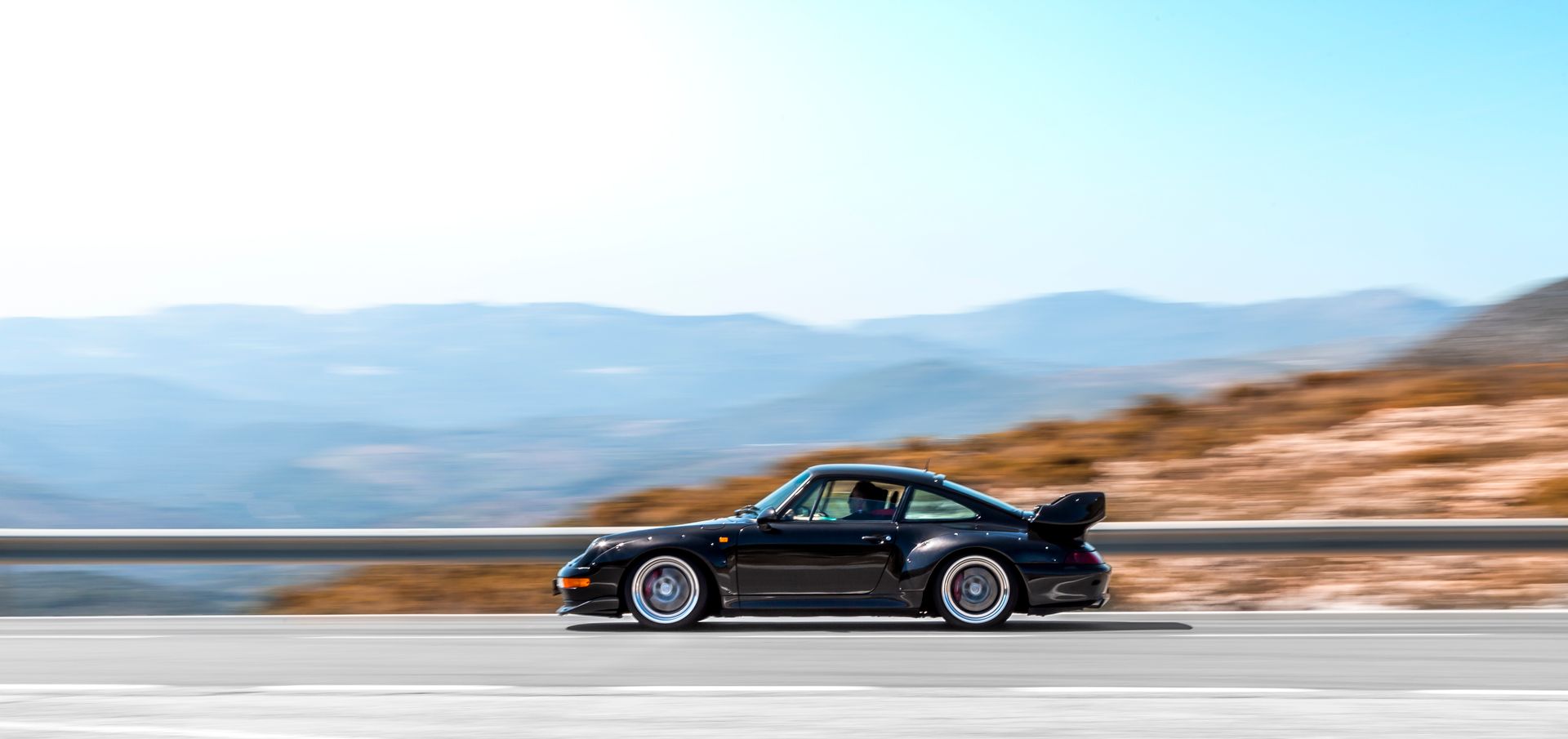 A sleek black supercar speeding on a curvy mountain road during bright daytime.