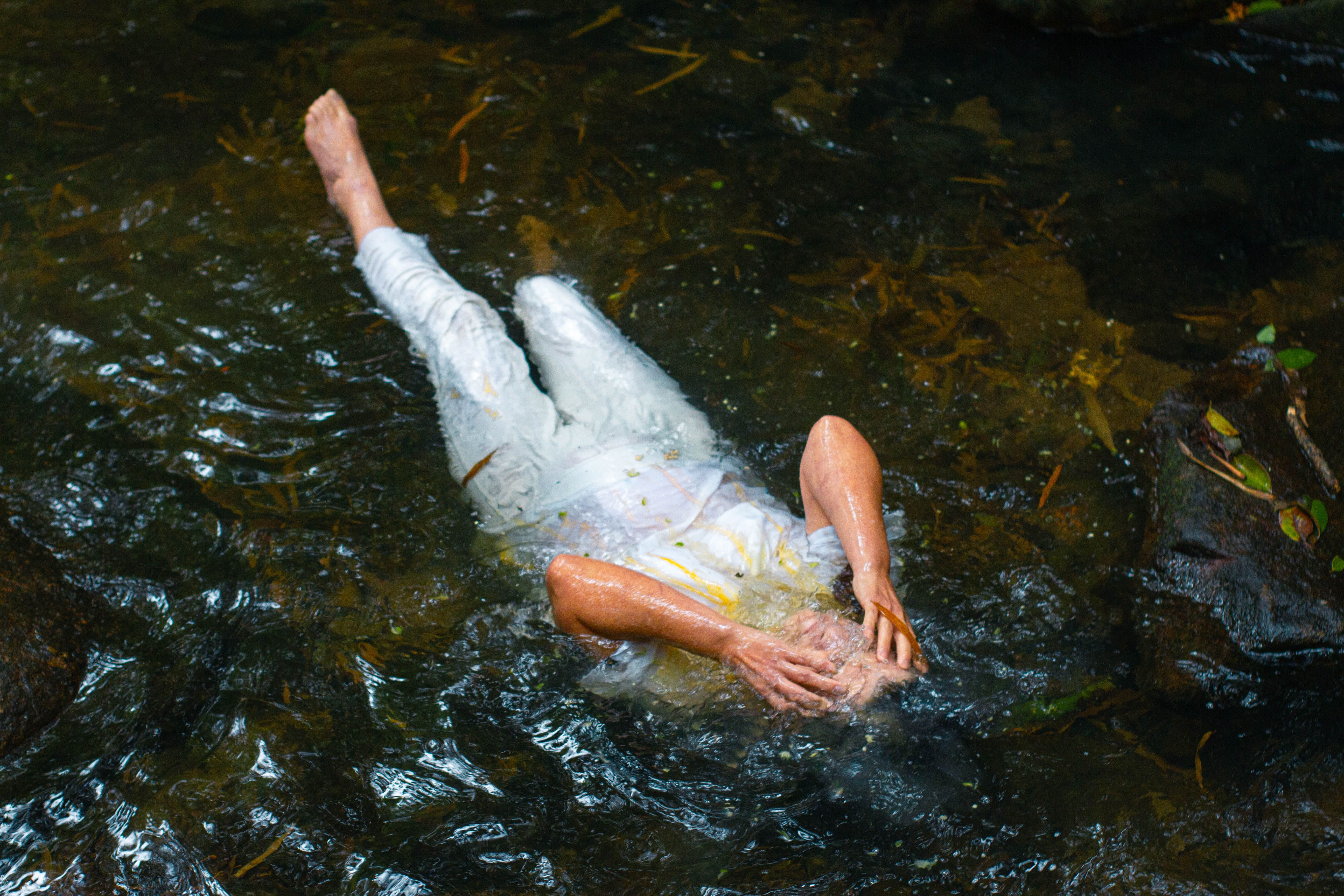 A person in white clothing lying down in a peaceful stream, surrounded by clear water.