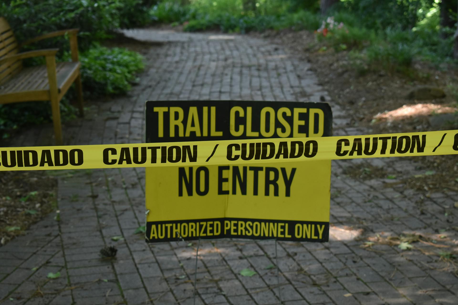 Trail closed sign with caution tape blocking pathway in a park setting.