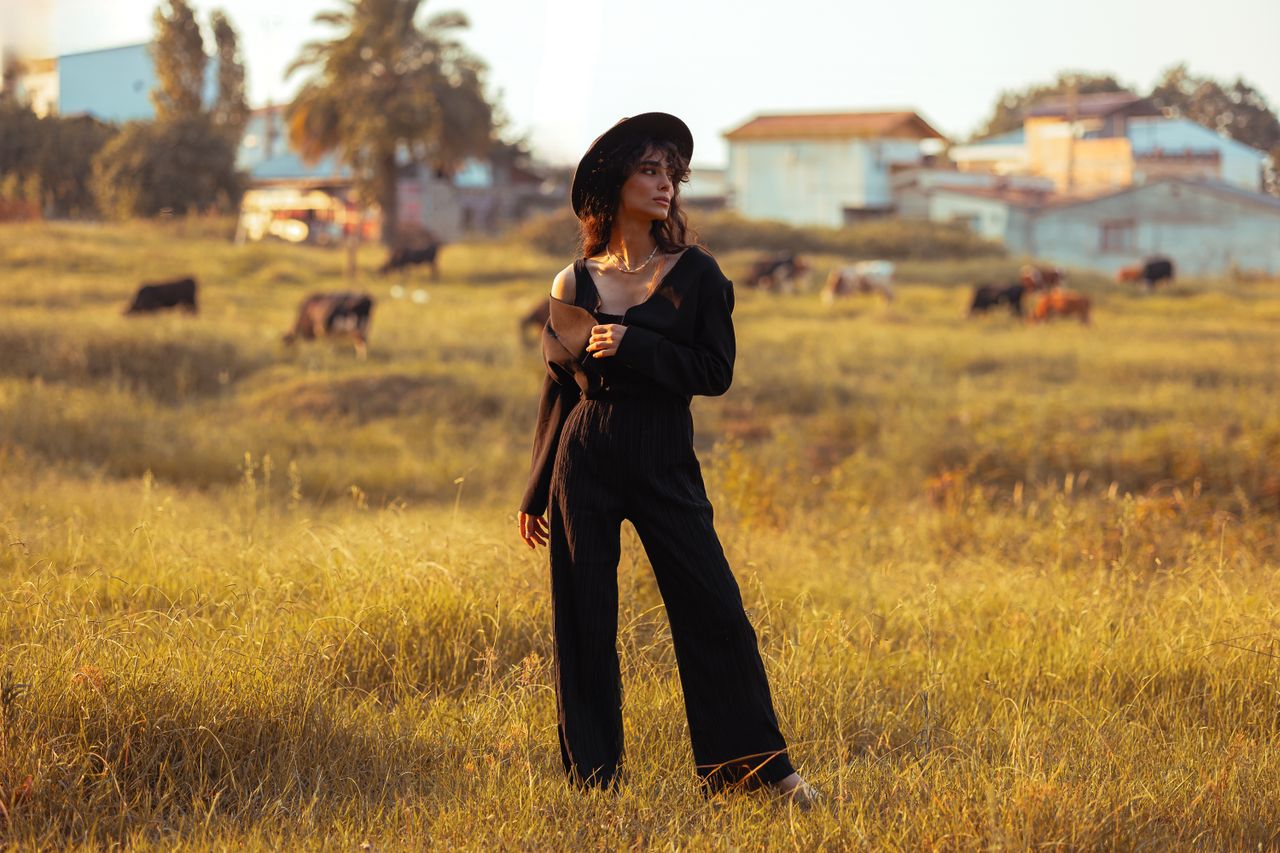 woman in black outfit and hat in a rural field with cows in the backgr