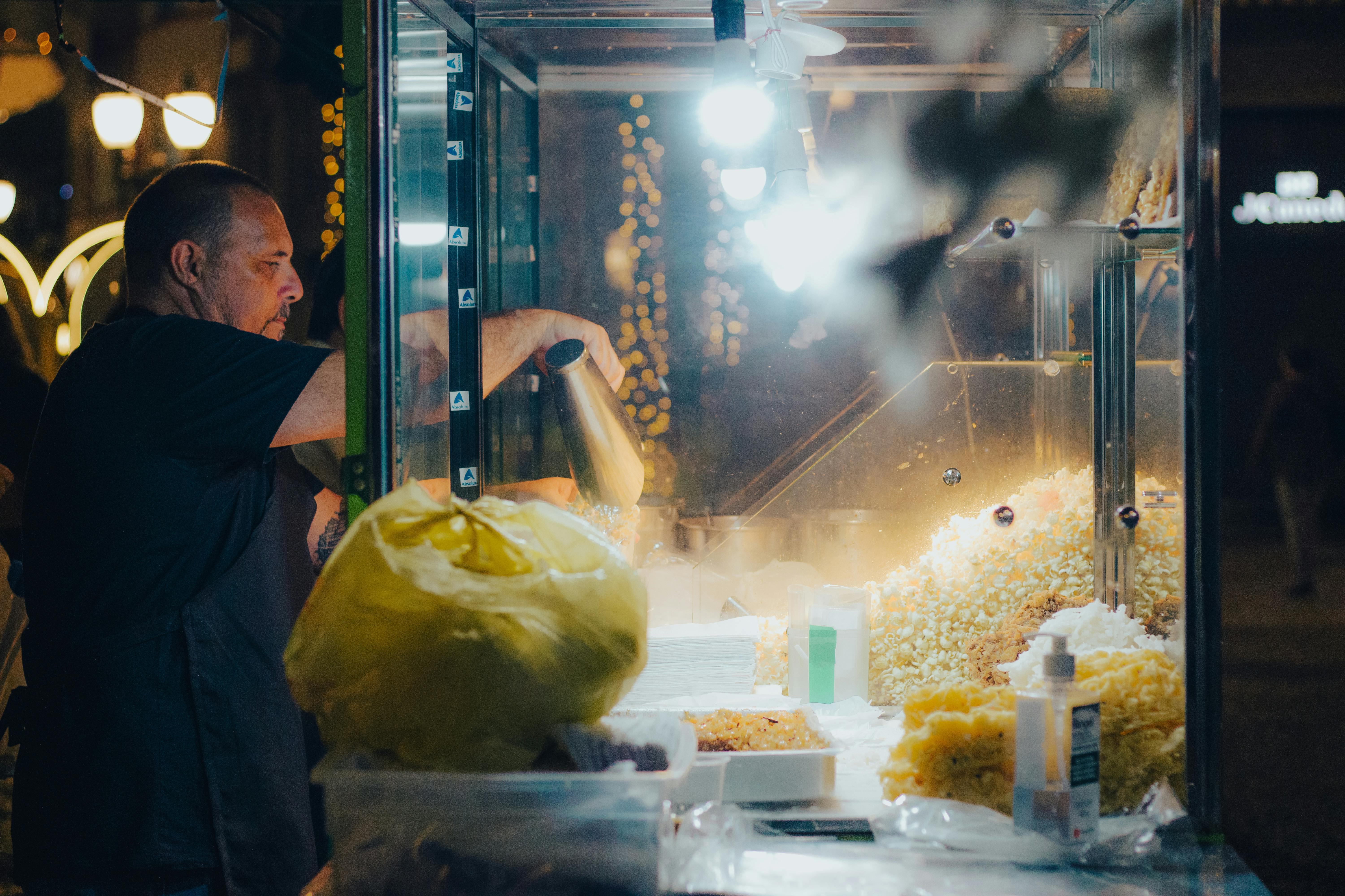 Gratuit Un vendeur ambulant prépare du pop-corn frais sur un stand de marché nocturne, éclairé par des lumières vives. Photos