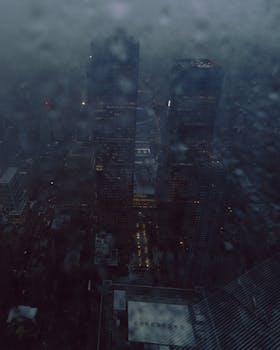 An aerial view of a cityscape at night, obscured by rain. Tall buildings are visible through the overcast sky.