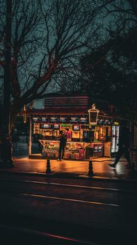 Cozy street food stall lit warmly at nighttime, inviting passersby. Urban ambiance with tree-lined path.