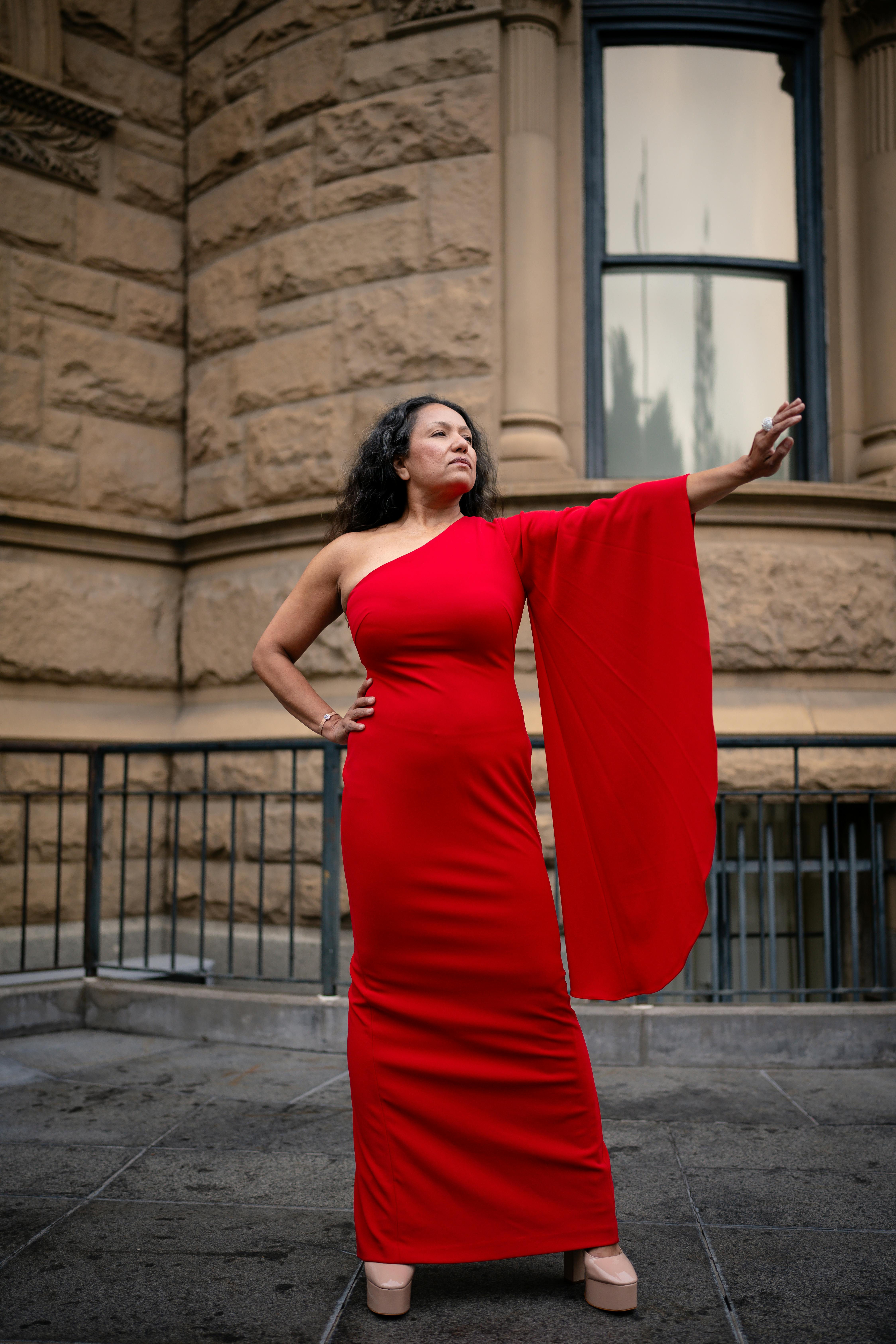 Stylish woman in a striking red dress poses confidently against a historic stone building facade.