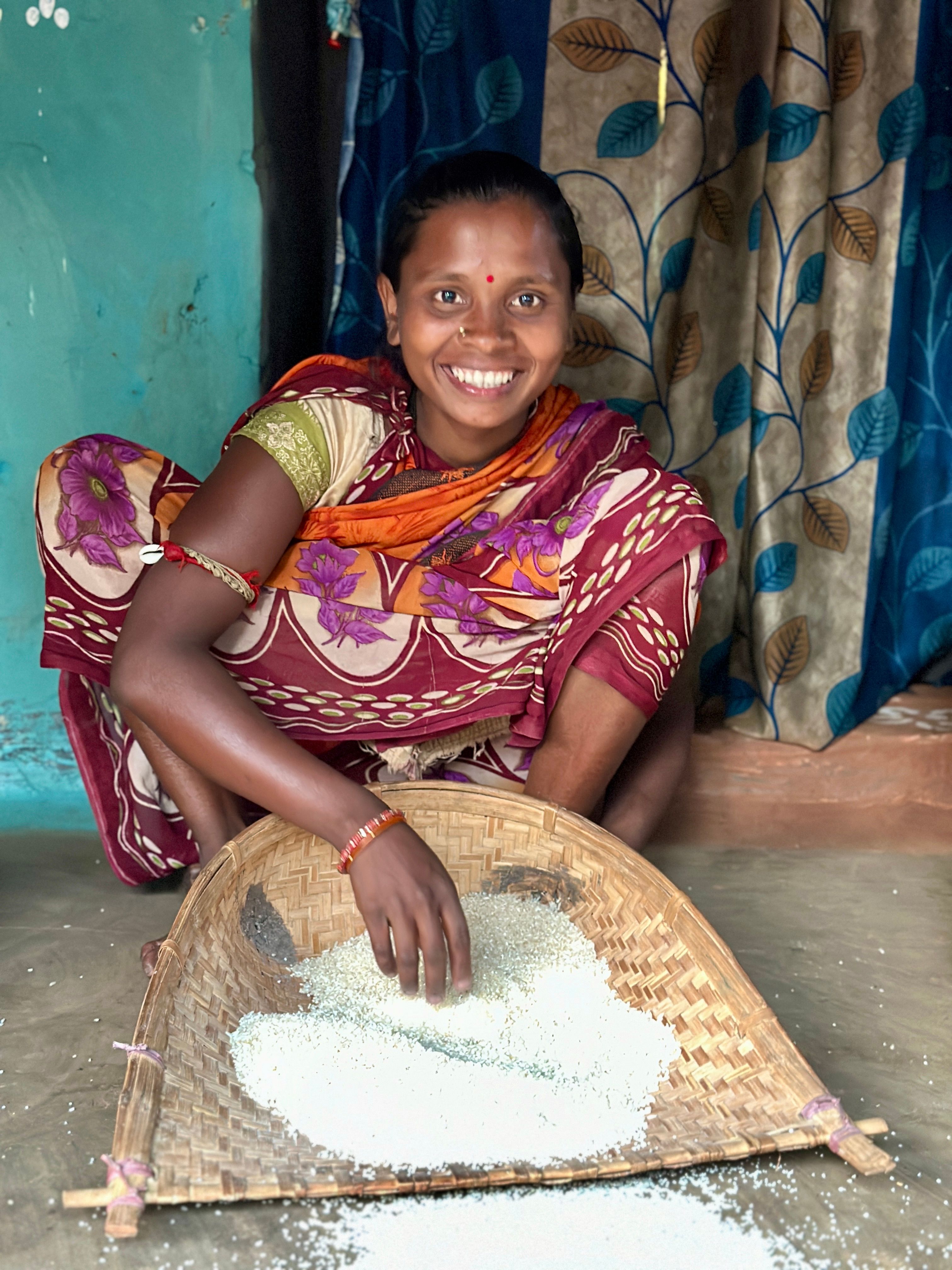 Smiling woman in colorful saree sifts grain using traditional method indoors.