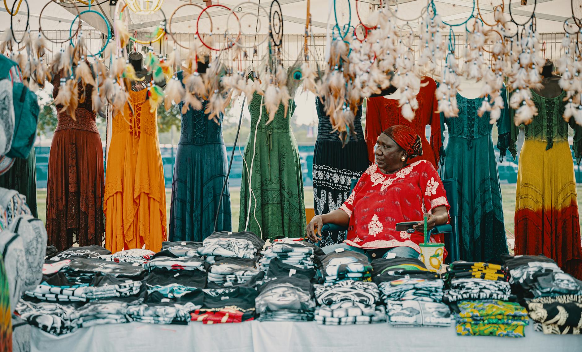 Colorful outdoor market stall featuring dresses, dreamcatchers, and a vendor offering diverse handmade items.