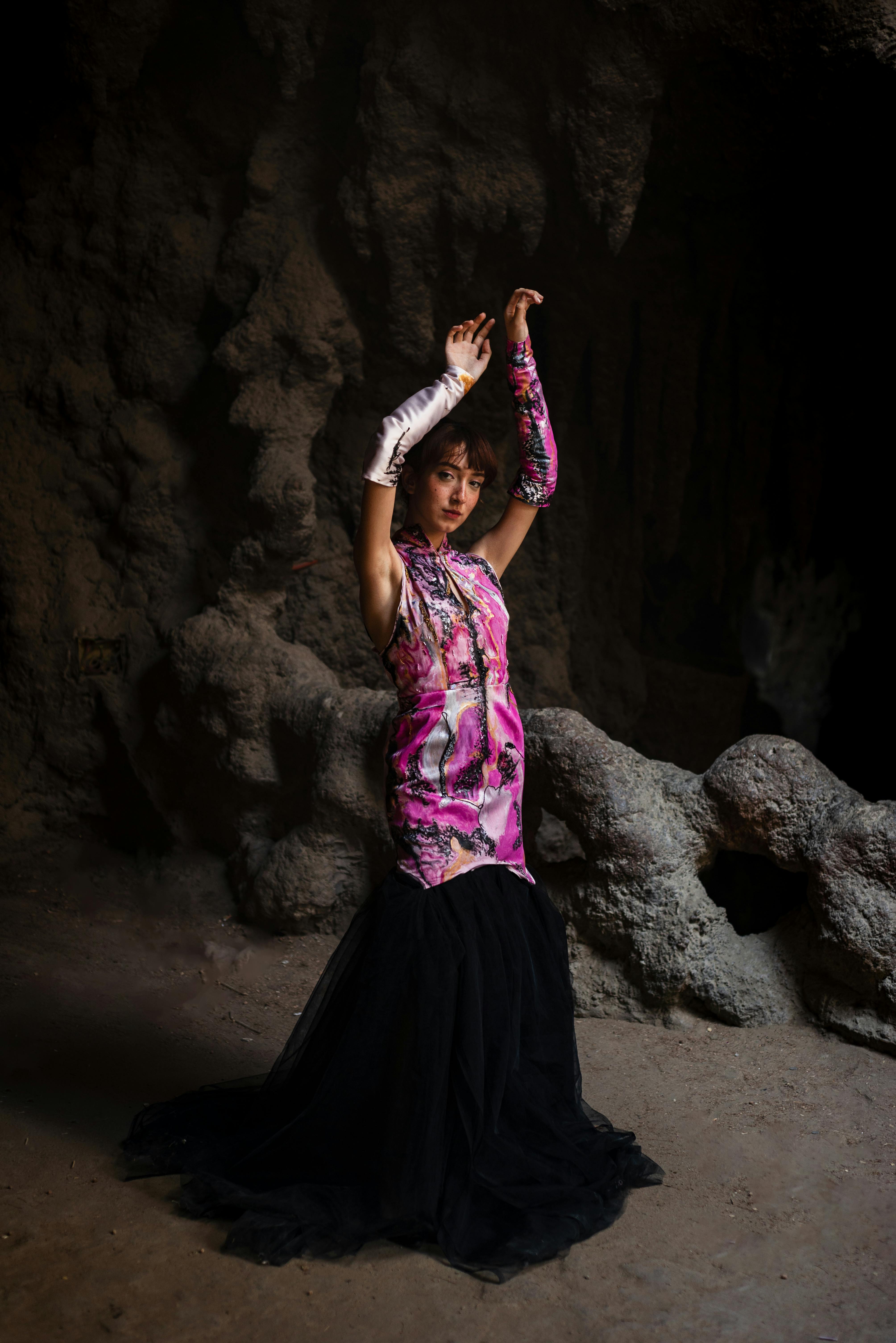 Woman in a pink dress poses confidently in a dark cave, exuding elegance and mystery.