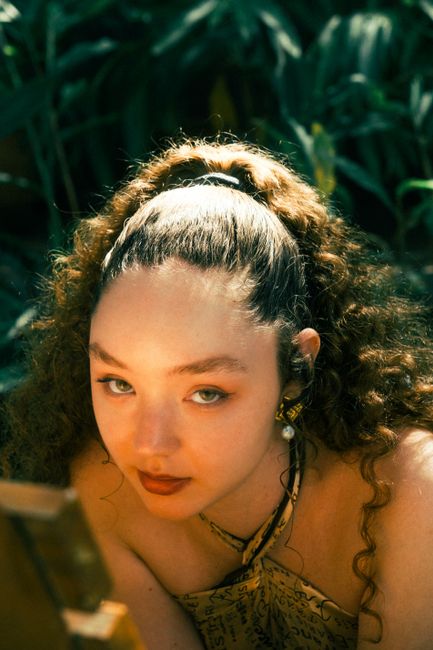 Close-up of a woman with curly hair in a sunlit, tropical setting, looking at the camera.