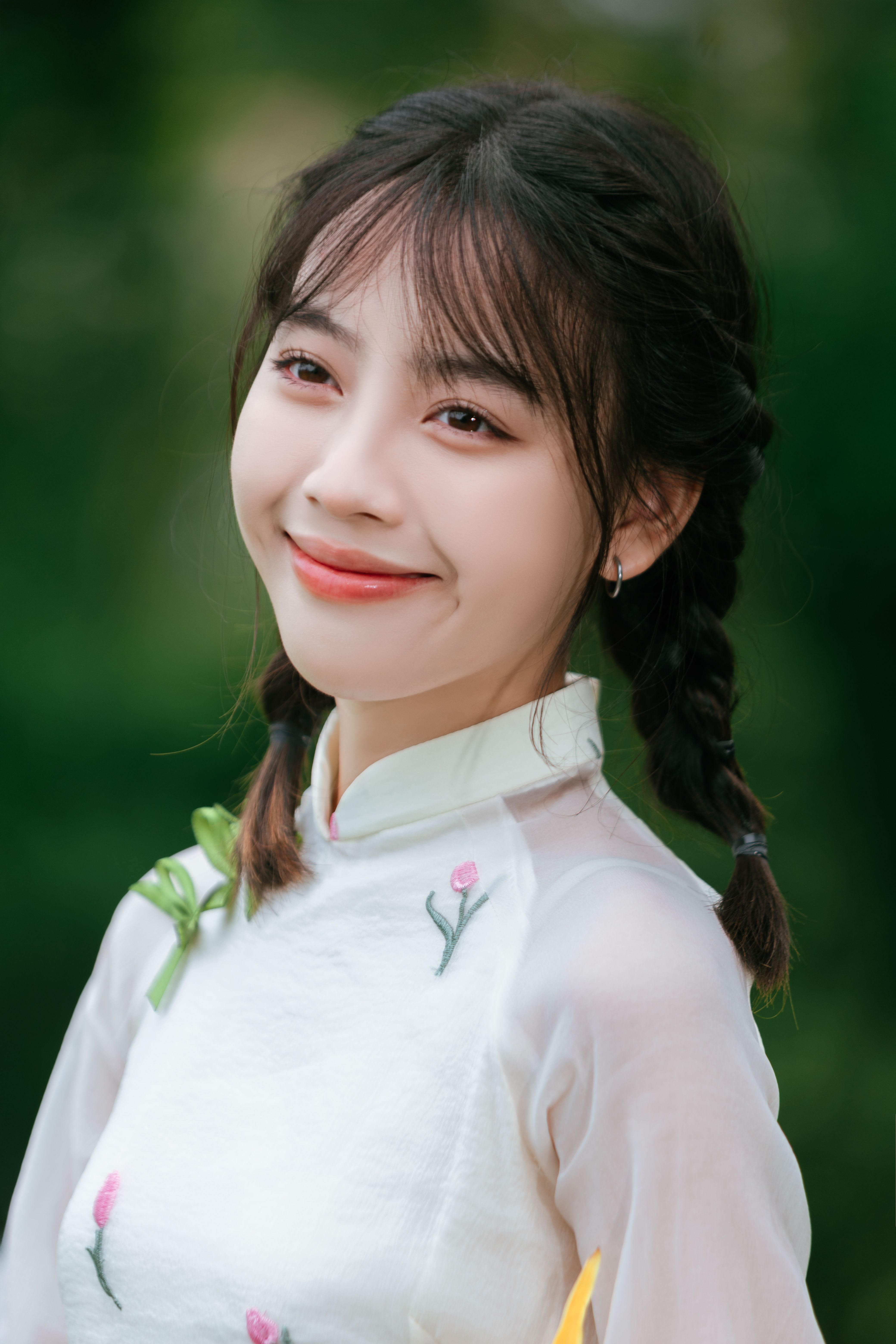 A young woman with braided hair and a white floral top smiles warmly against a blurred green backgro
