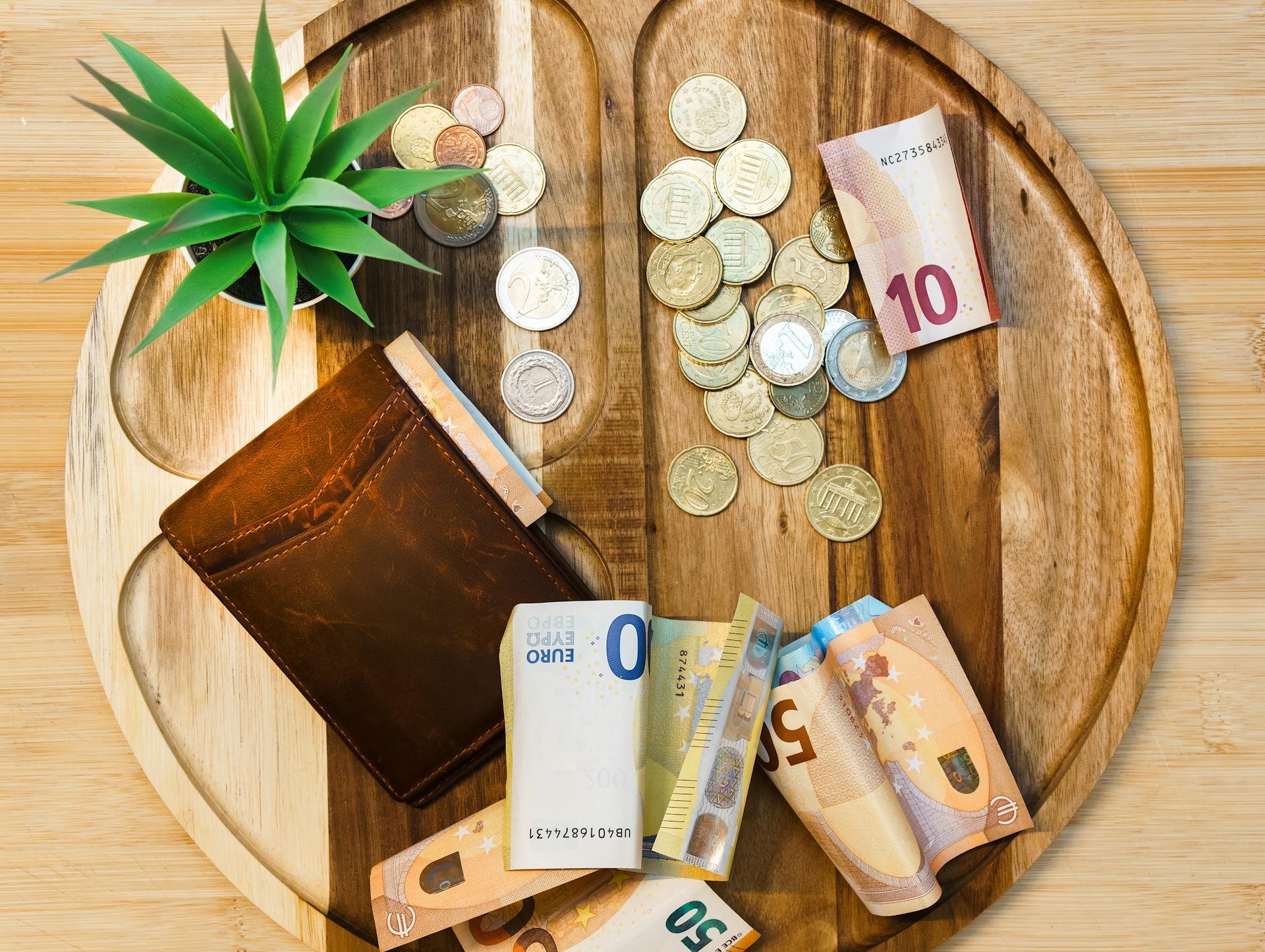 Euro banknotes and coins on a wooden tray with a wallet and plant.