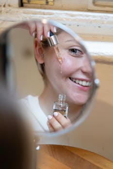 Smiling woman applying face serum with dropper seen in mirror reflection.