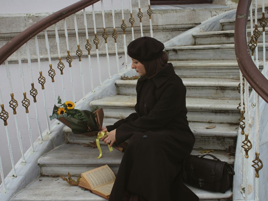 Woman in black coat and beret sitting on stairs with sunflowers and open book.