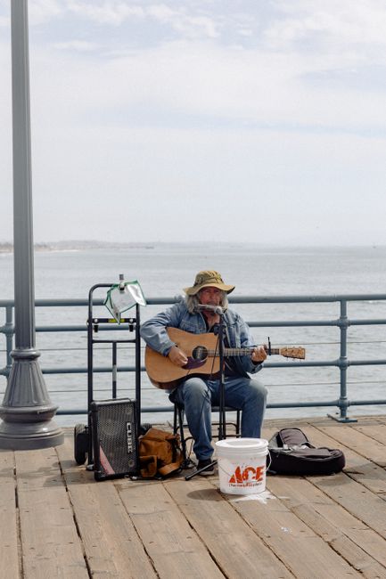 Senior man plays guitar by the ocean on a sunny day, showcasing street music culture.