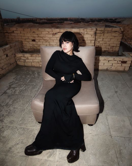 Elegant woman in black dress sitting on a rooftop chair with brick wall backdrop.