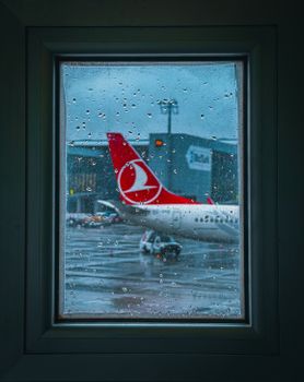 View of aircraft and terminal through raindrop-covered window, reflecting travel mood.