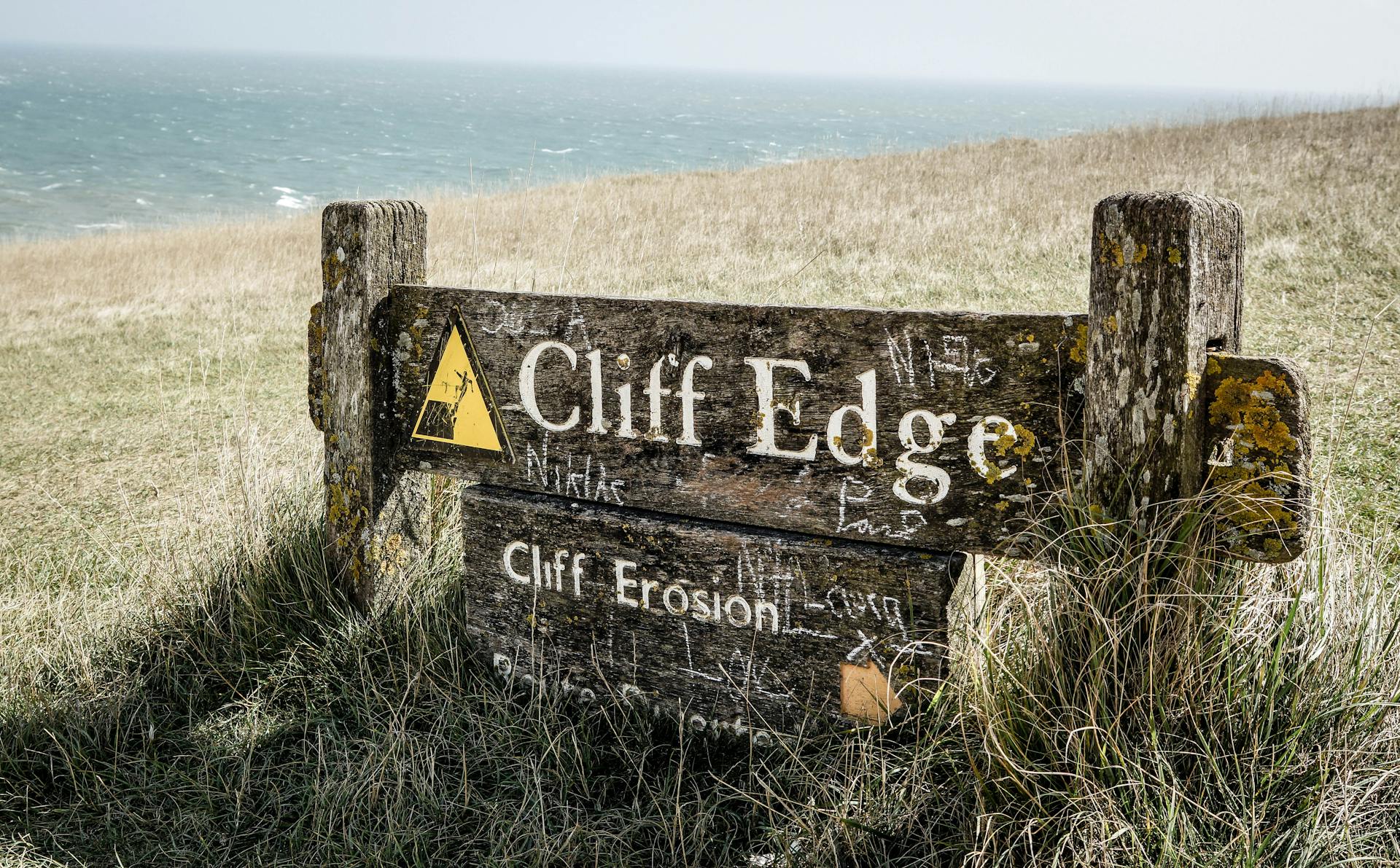 Weathered warning sign at a coastal cliff edge highlighting erosion risk with an ocean backdrop.