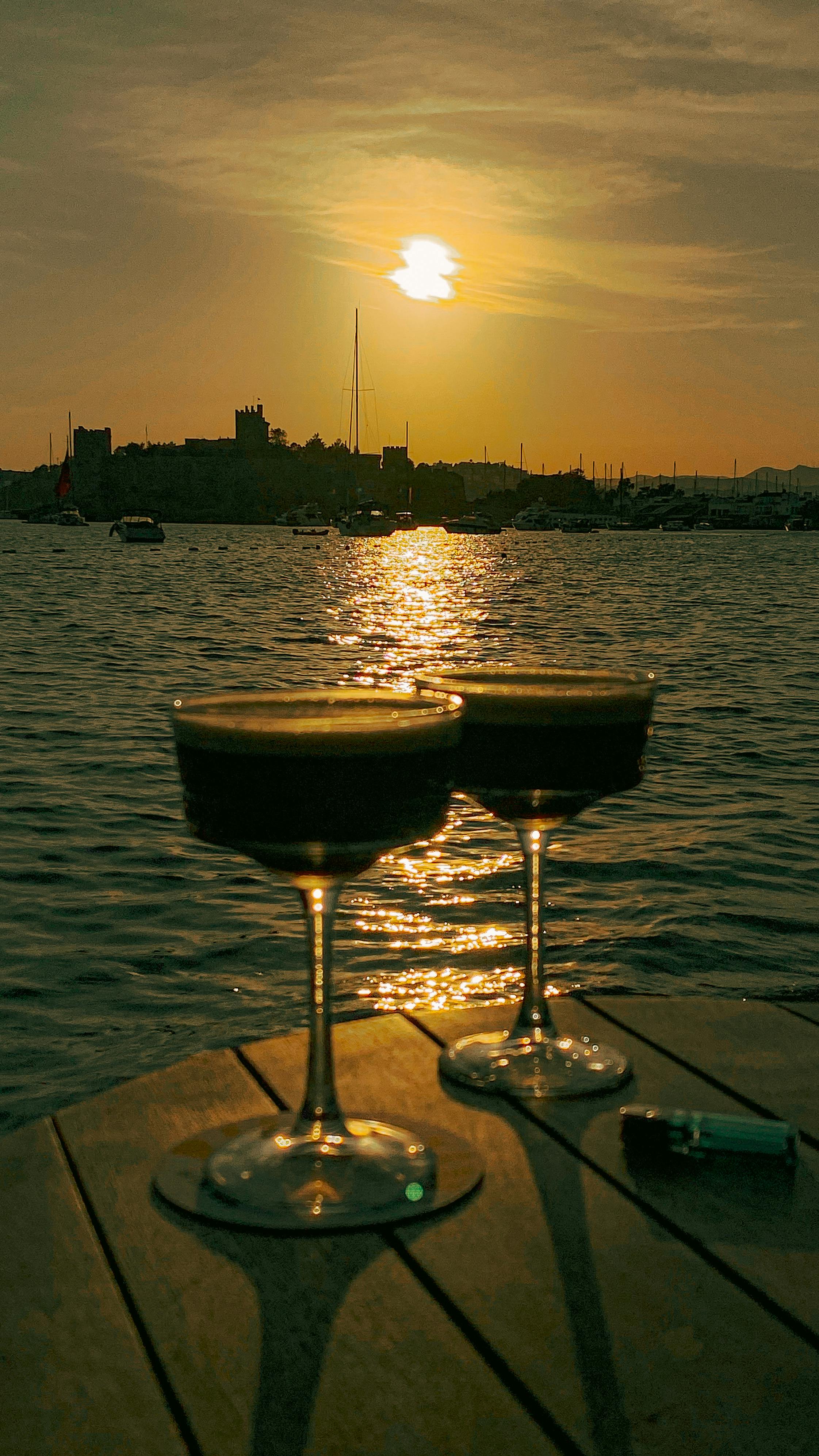 Elegant cocktail glasses on a table by the water, with a sunset and castle silhouette backdrop.